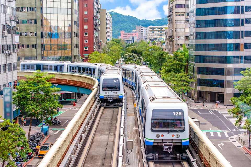 Two modern metro trains on elevated tracks in a cityscape with tall buildings, green trees, and a mountainous backdrop.