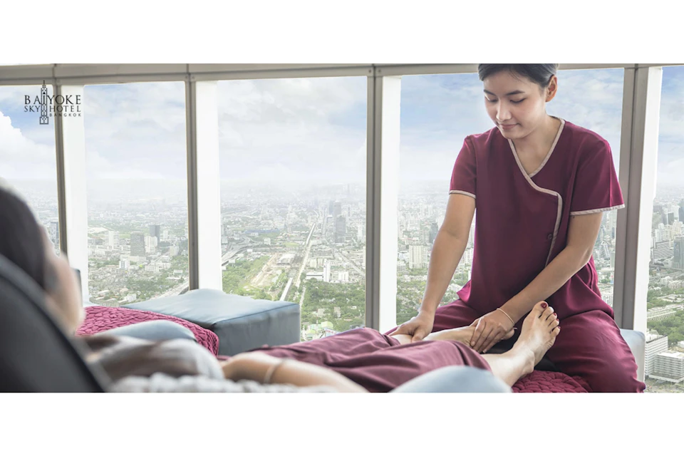 A massage therapist in a red uniform works on a client's foot in a high-rise setting with a cityscape view through large windows.