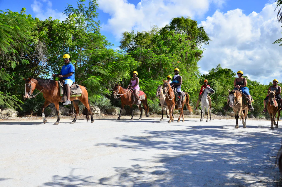 People wearing helmets riding horses on a dirt path, surrounded by greenery under a partly cloudy sky.
