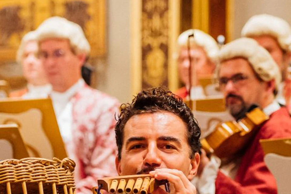 Musicians in baroque-inspired costumes perform with period instruments on stage, one man in colorful attire plays a pan flute.