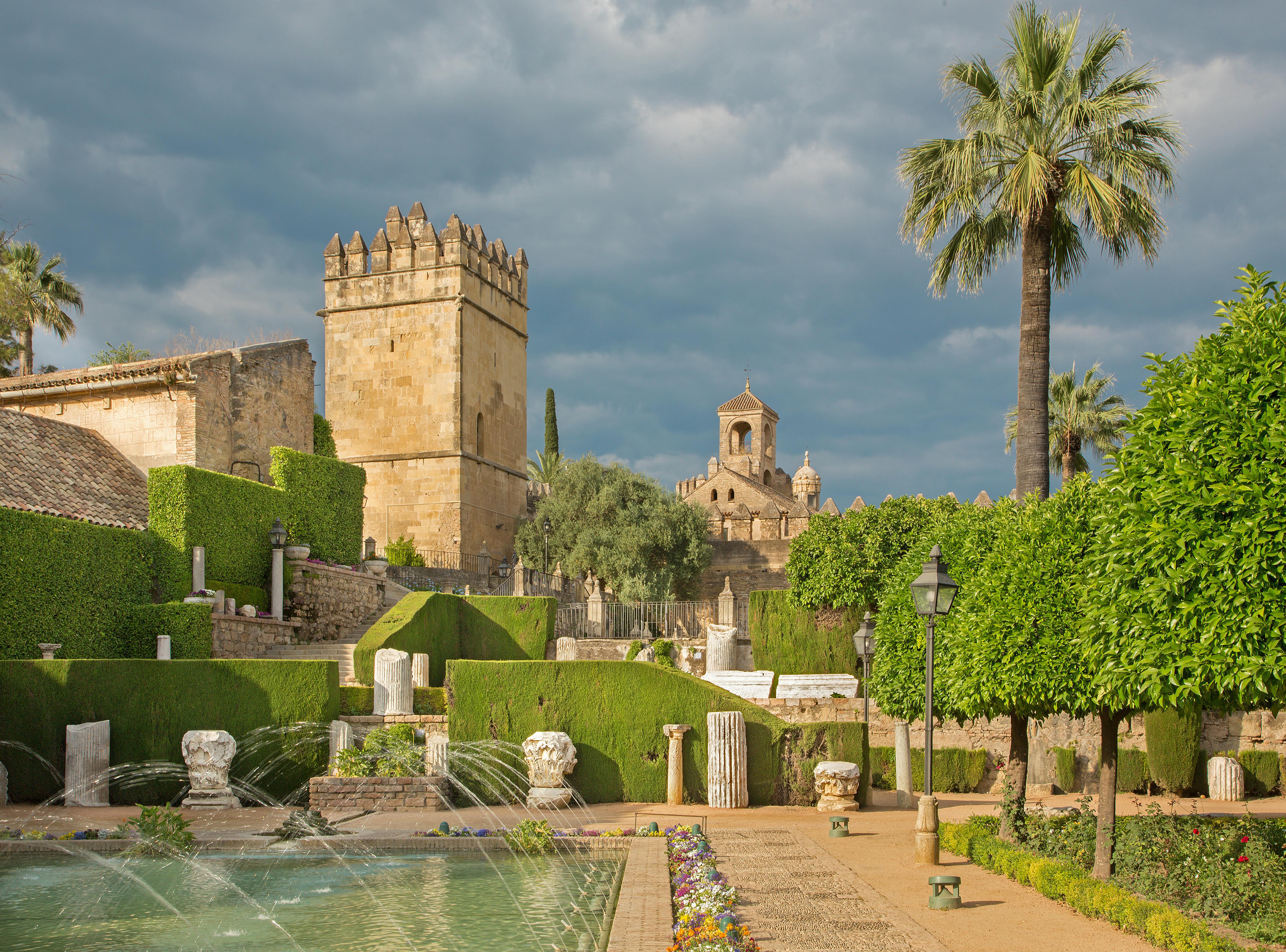 Ancient stone tower and church surrounded by lush gardens, fountains, and palm trees under a cloudy sky.