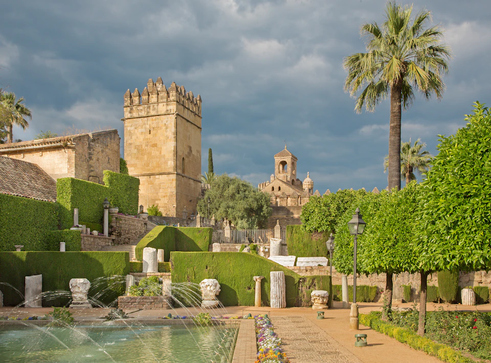 Ancient stone tower and church surrounded by lush gardens, fountains, and palm trees under a cloudy sky.
