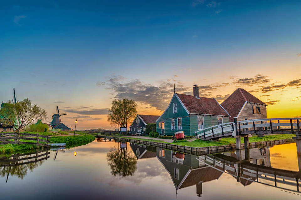 A serene canal scene at sunset with quaint houses, a bridge, tree, and a windmill reflecting on the water.