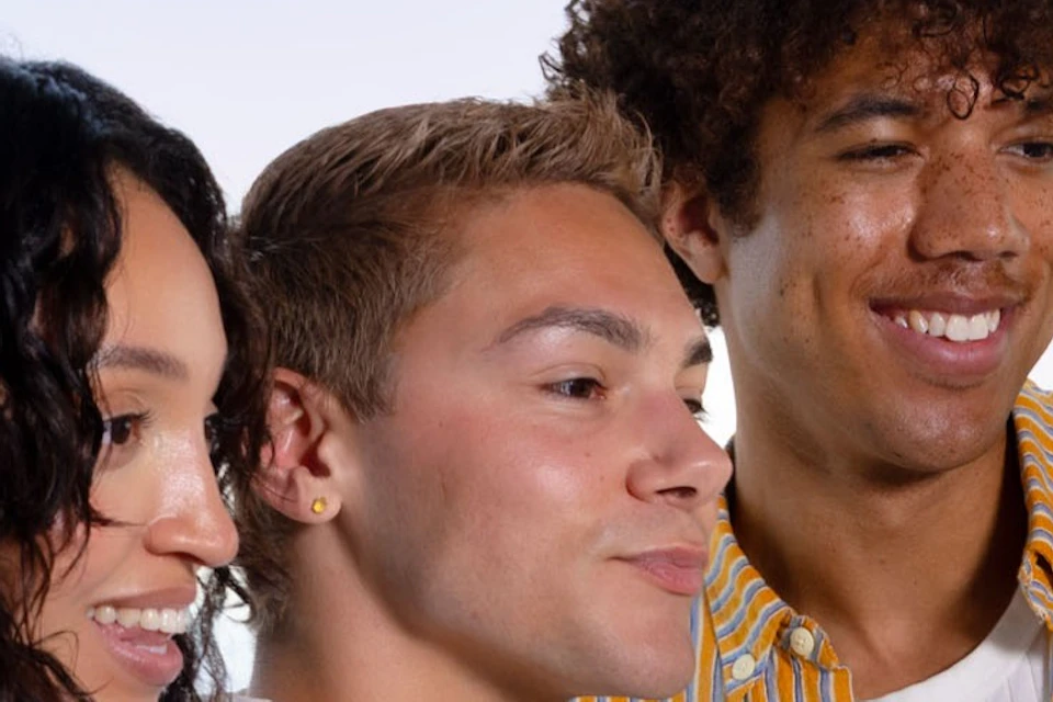 Three people taking a group selfie near a large window with a cityscape and blue sky visible in the background.