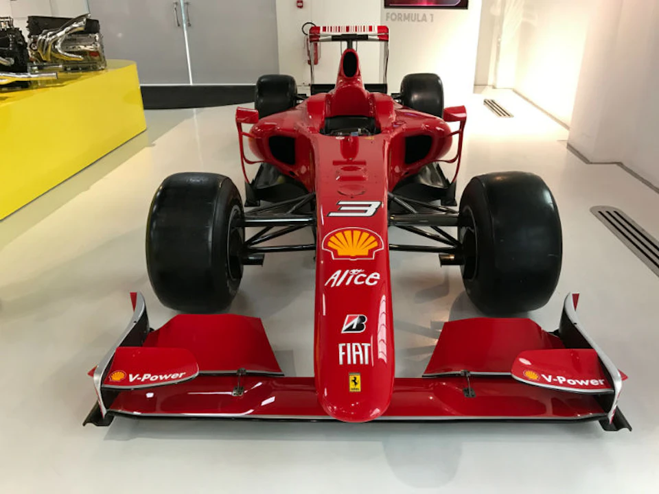 Red Formula 1 racing car with various sponsor logos, displayed indoors on a glossy white floor.