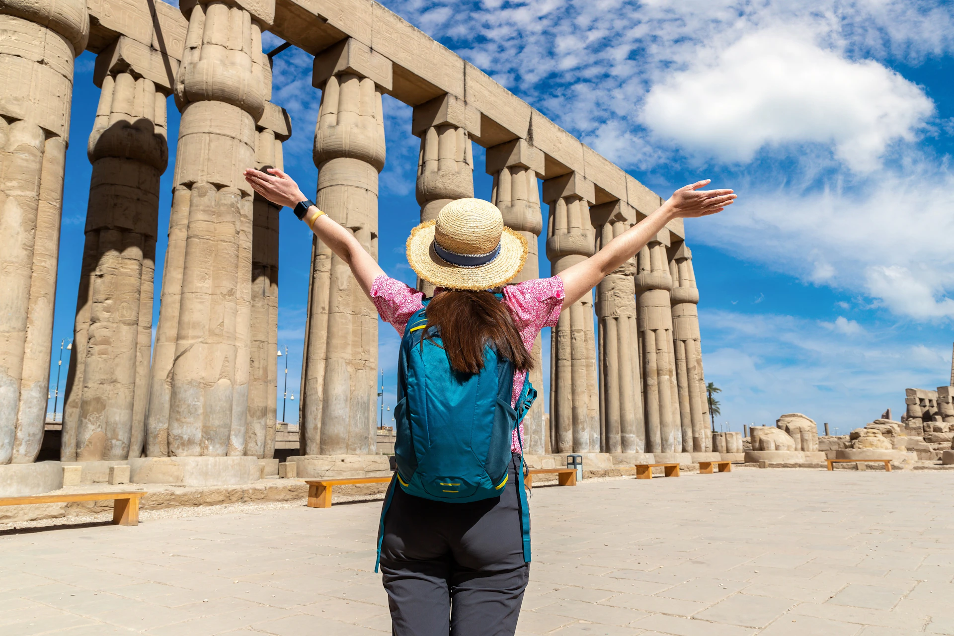 A person wearing a hat and backpack raises their arms in front of ancient stone columns under a partly cloudy sky.