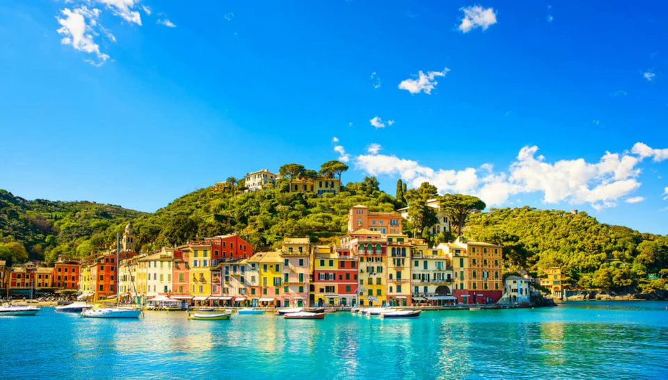 Colorful buildings line a waterfront, with boats docked in front and a hillside with greenery and more buildings in the background.
