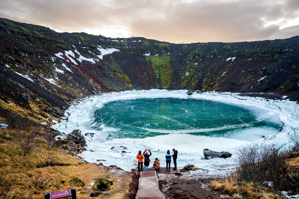 A group of people standing at the bottom of the crater in front of the lake in winter. Moss and red rocks