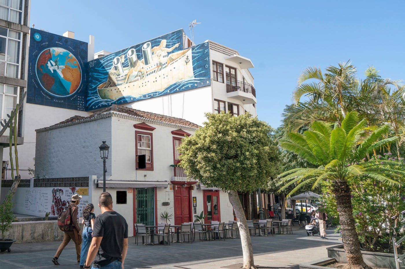 La gente pasa ante un edificio con murales de colores, asientos al aire libre y exuberante vegetación bajo un cielo azul despejado.