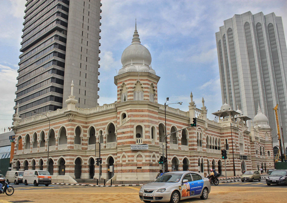 A historic building with arched windows and domes is flanked by modern skyscrapers. Cars and motorcycles pass by.