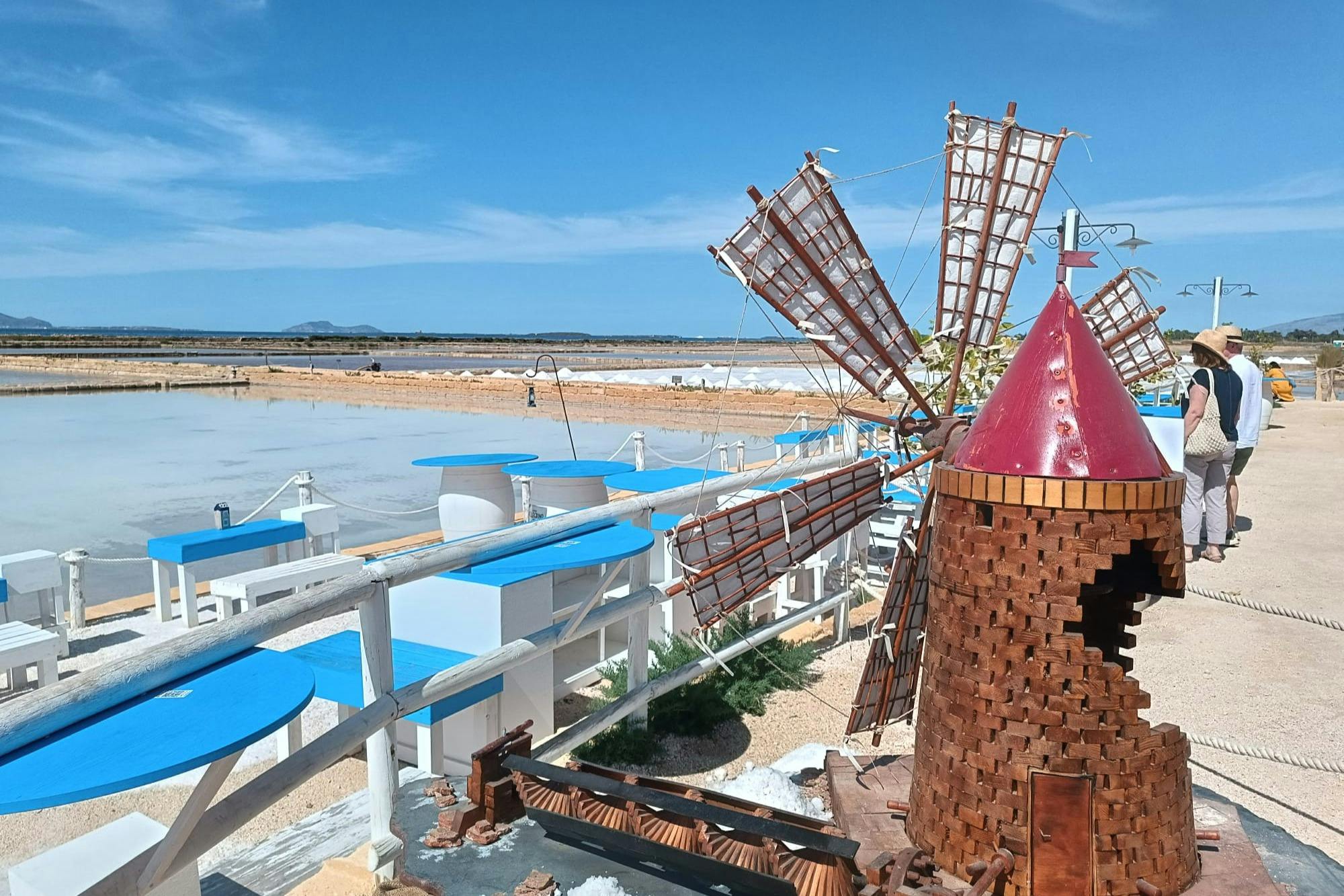 A rustic windmill beside a series of salt pans under a clear blue sky, with white and blue structures in the foreground.