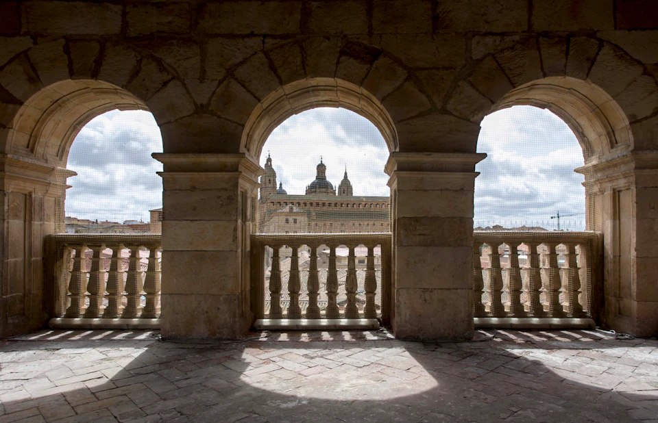 Stone archways frame a view of a historic building with domes and towers under a cloudy sky. Shadows fall on a tiled floor.
