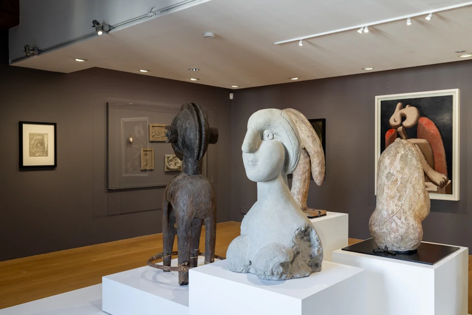 A museum display featuring abstract sculptures on white pedestals, with framed art on the walls in a modern gallery setting.