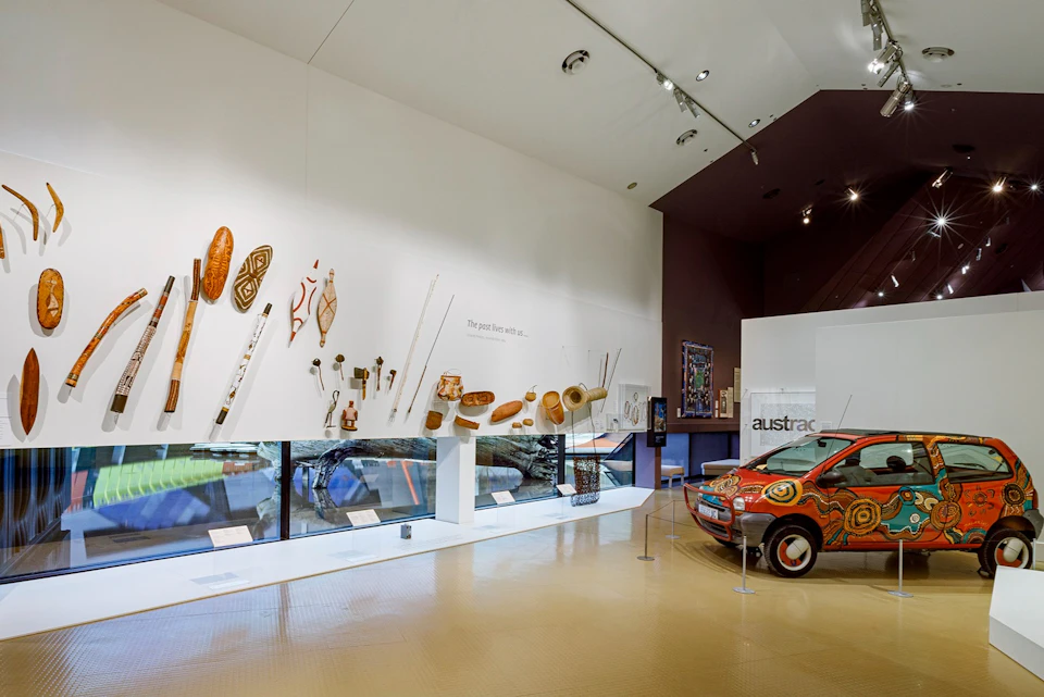 Museum exhibit featuring Indigenous art, artifacts, and a decorated car, displayed in a modern, well-lit gallery space.