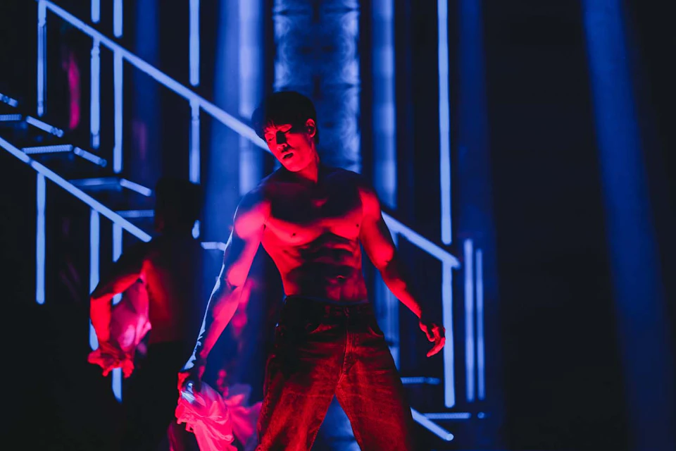 A shirtless person stands under dramatic red lighting, surrounded by a dark blue background with neon-like geometrical designs.