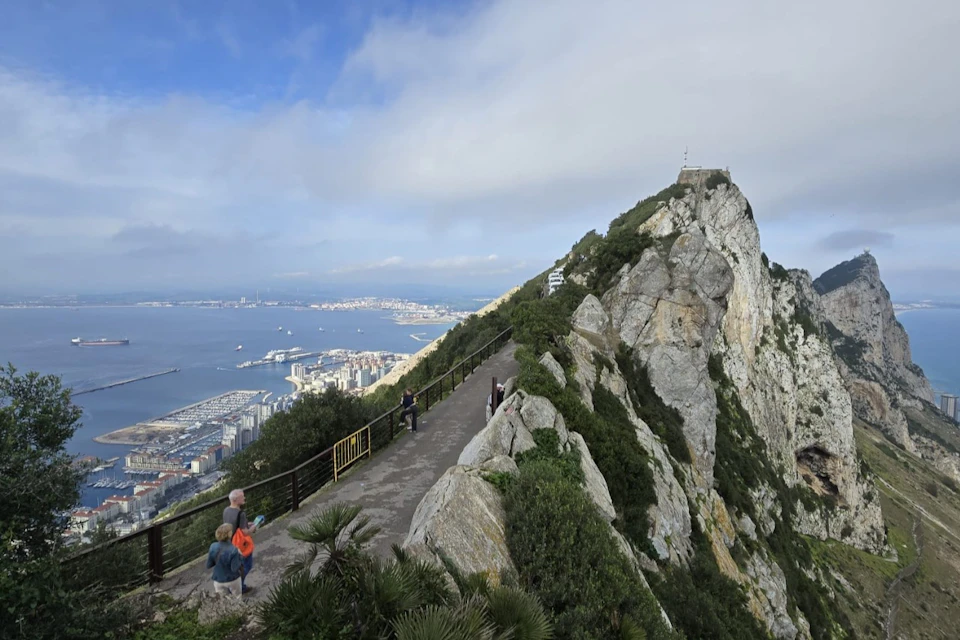 A footpath along a ridge offering views of a city, harbour and the sea below, surrounded by lush greenery and rocky terrain.