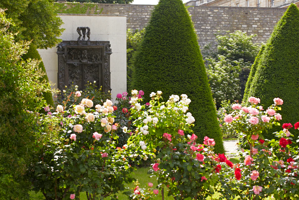 Garden with blooming pink, red, and white roses, manicured conical trees, and a stone wall with a dark ornate relief.