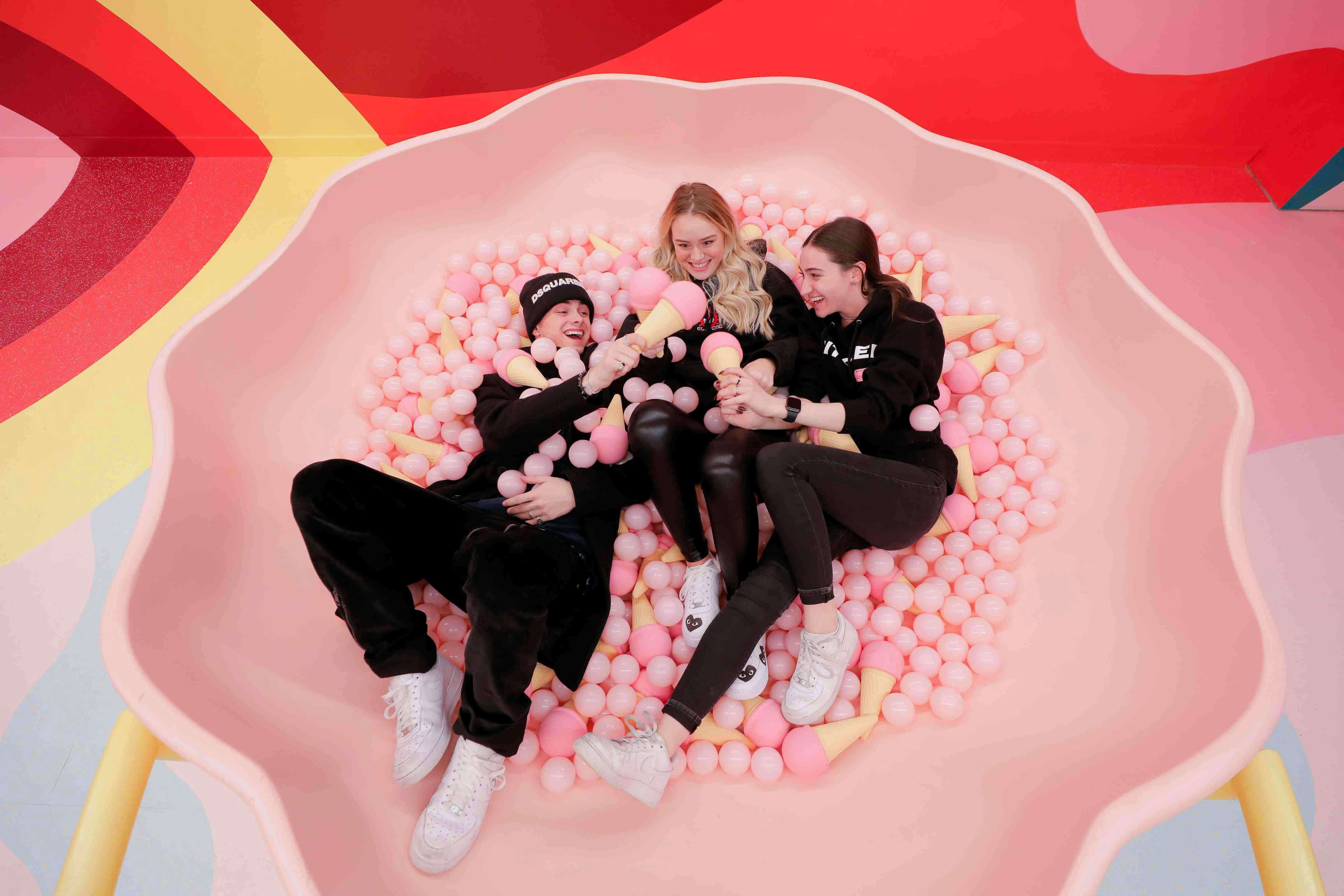 Tři lidé sedí v růžové míčové jámě, drží zmrzlinové kornouty, usmívají se a vzájemně se ovlivňují.