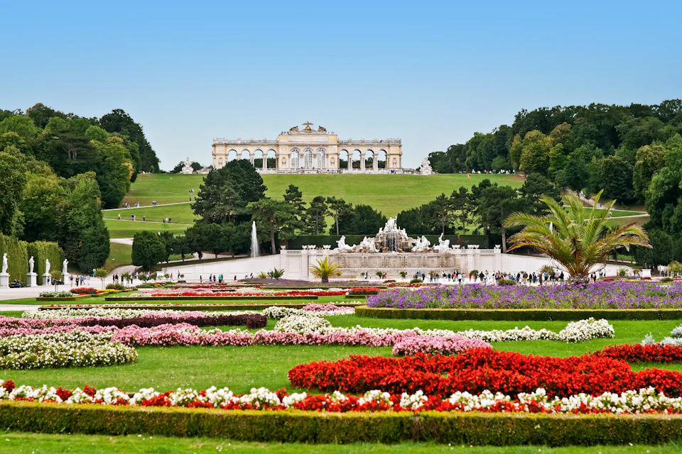 Vibrant garden with colorful flower beds, a central fountain, and a grand historic building on a hill under a clear blue sky.