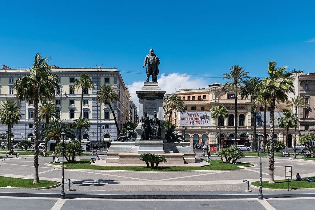 晴朗的蓝天下，公共广场上有雕像、棕榈树、人行道和历史建筑。