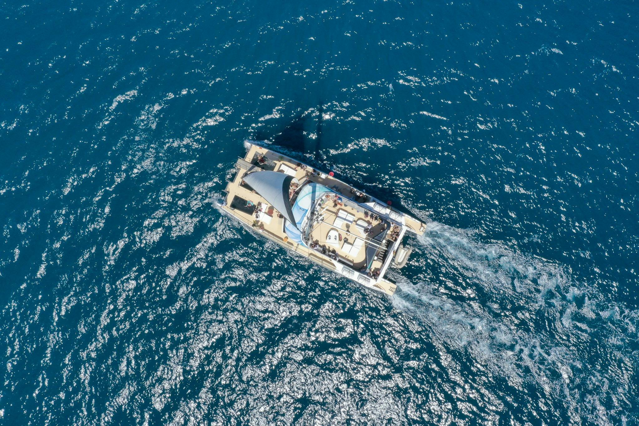 Luftaufnahme eines Katamarans, der auf einem blauen Meer segelt, mit sichtbarem Kielwasser und Menschen an Deck.