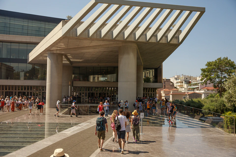 People walk towards a large, modern building with a slatted roof and glass facade under a clear sky.