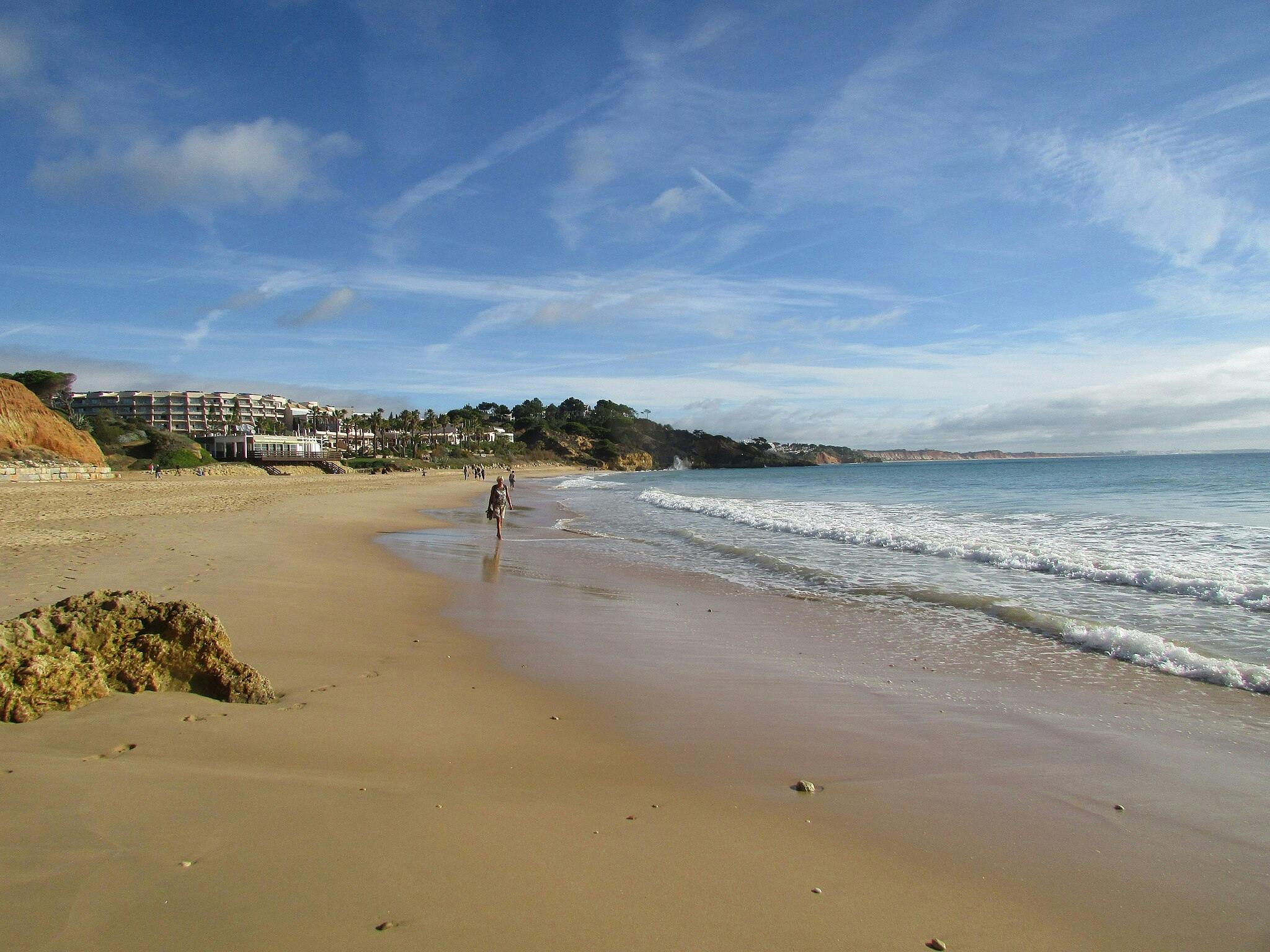 Písečná pláž s jemnými vlnami, pár lidí kráčejících po břehu, útesy a budovy v pozadí, modrá obloha s jemnými mraky.