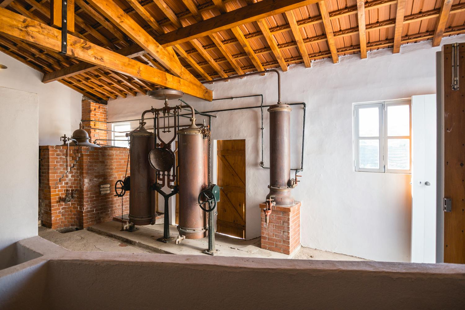 Interieur van een rustieke distilleerderij met koperen en metalen apparatuur tegen witte muren en een houten plafond.