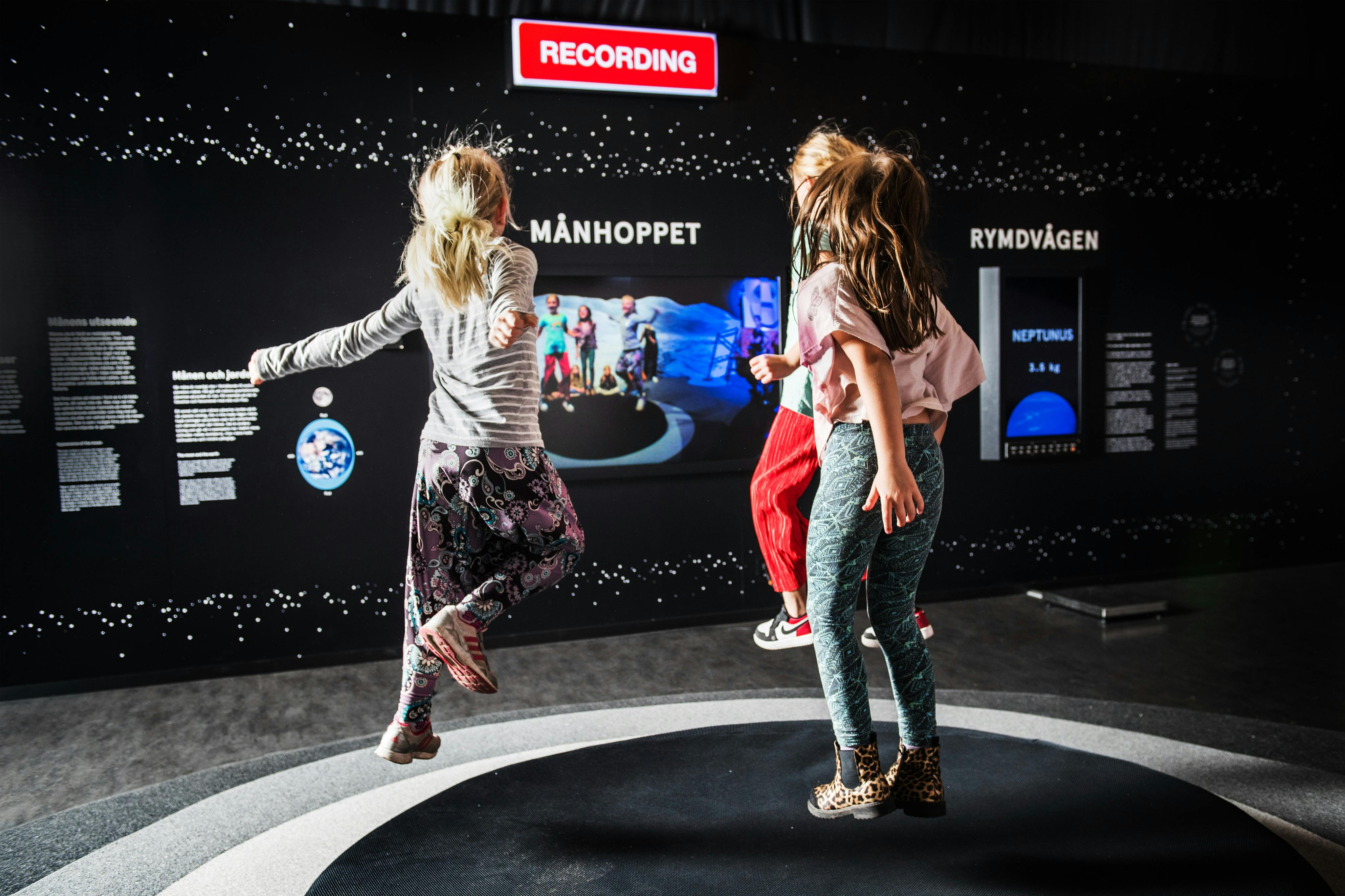 Três crianças a saltar e a brincar numa superfície circular numa área de exposição com o tema da lua, sob um sinal de "GRAVAÇÃO".