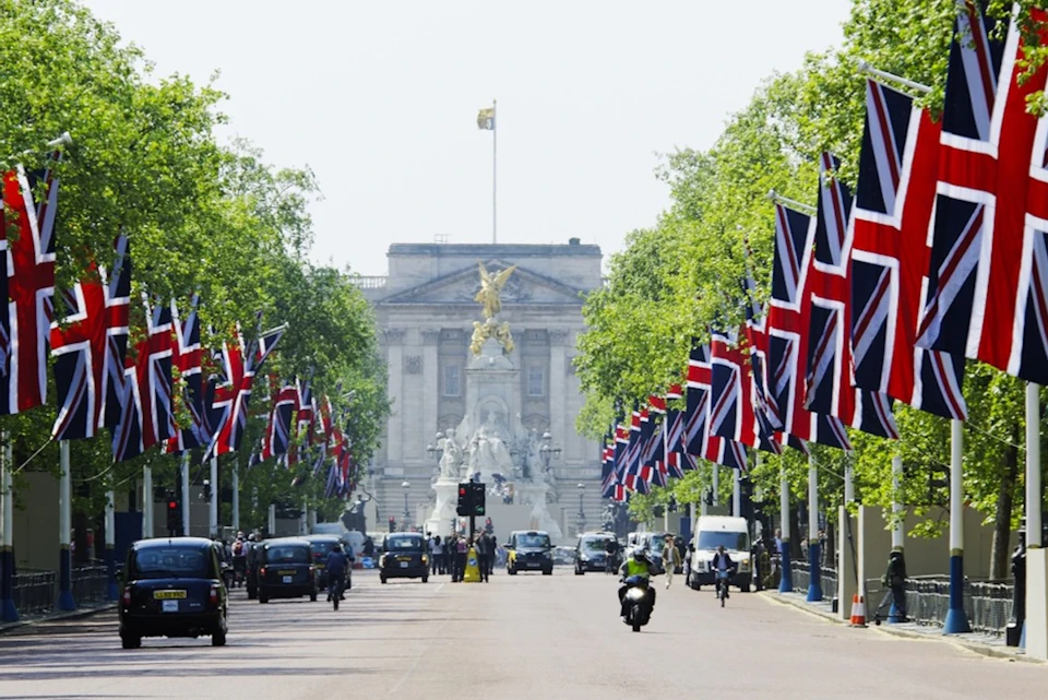 Buckingham Palace with its iconic gates, Union Jack flags lining the street, and traffic including taxis and a motorbike.