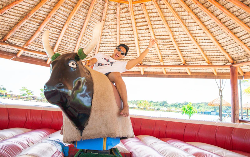 Person riding a mechanical bull indoors under a thatched roof, smiling and posing with one hand raised in the air.