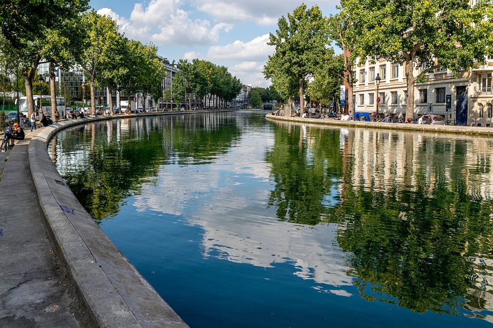 A calm canal lined with trees and buildings reflects a partly cloudy sky. People are walking and sitting along the sides.