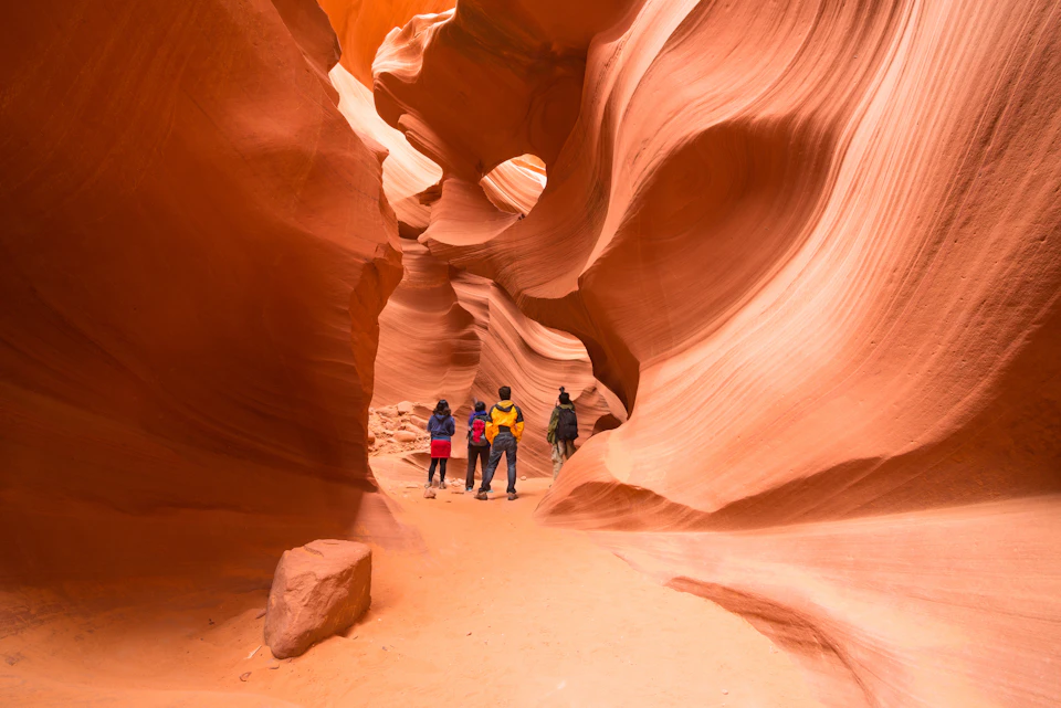 Four people stand in a narrow, winding sandstone canyon with smooth, flowing walls in shades of red and orange.