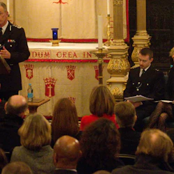 Mannen i uniform talar vid podiet framför den sittande publiken. En man och en kvinna sitter bredvid honom under ett dekorerat altare.