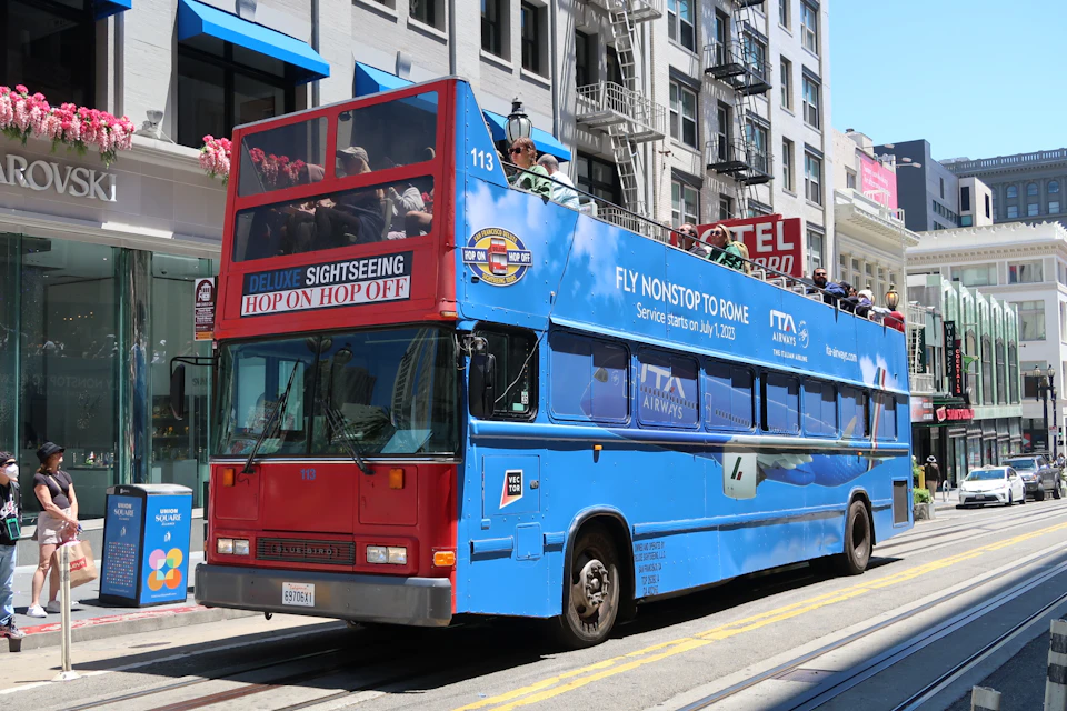Open-top blue sightseeing bus advertising flights to Rome, parked on a city street with tourists on board and buildings in the background.