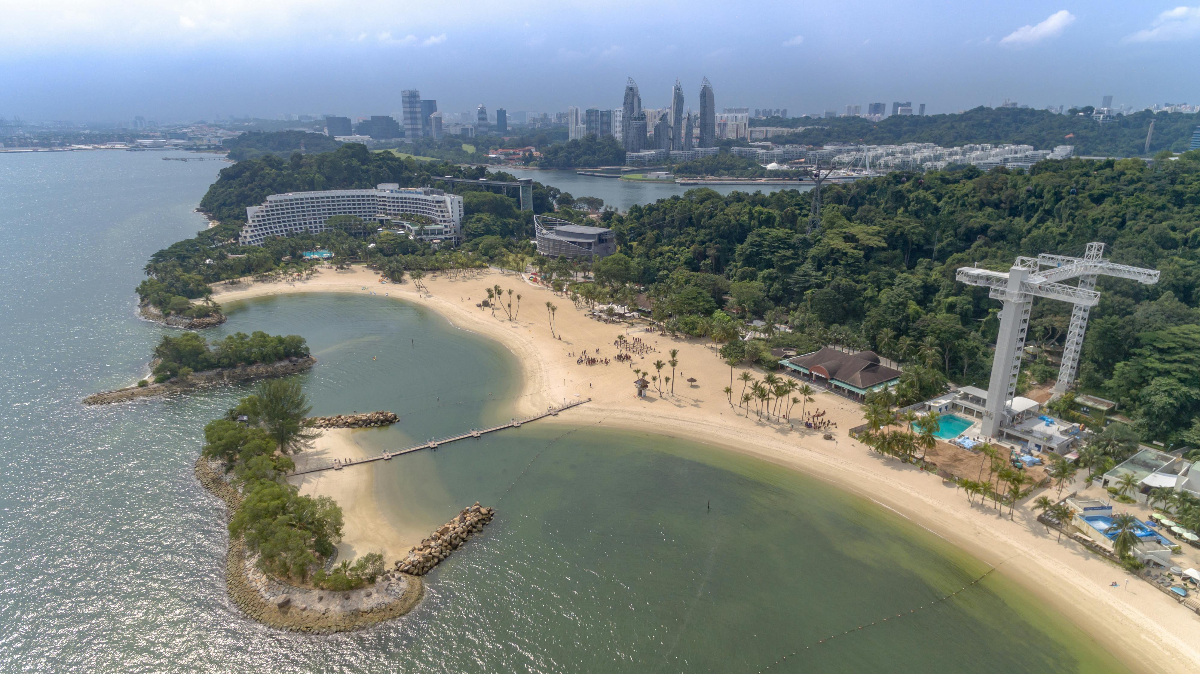 Aerial view of a beach with palm trees, a lagoon, and surrounding greenery. Cityscape with tall buildings in the background.