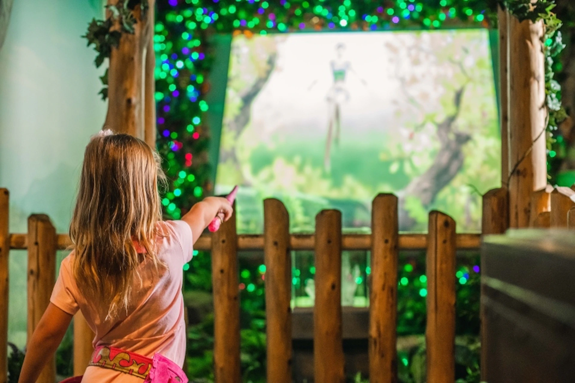 A child points towards a fairy-themed display screen in a garden decorated with colorful lights and wooden fencing.