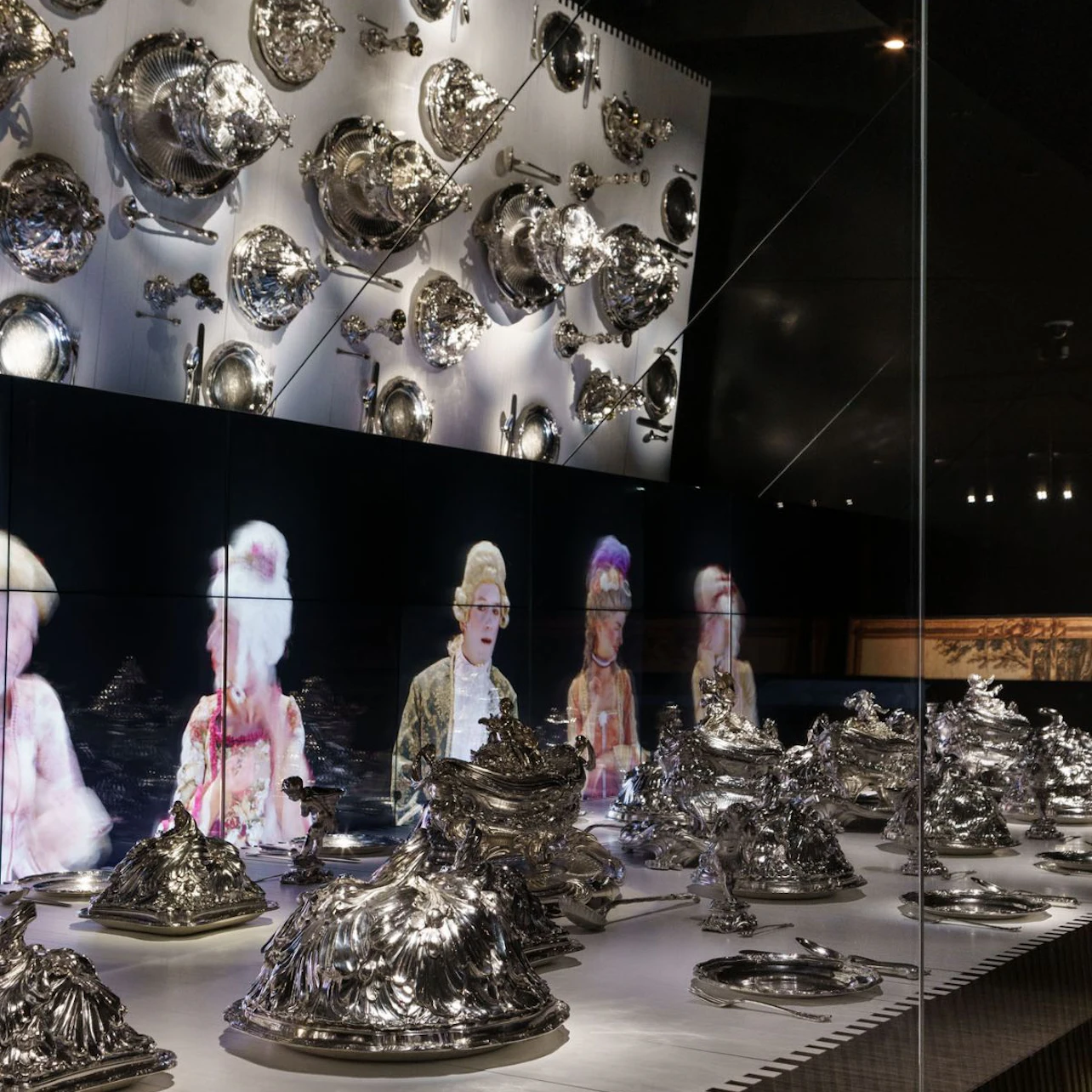Display case with an assortment of silverware on shelves and walls, and holograms of people in historical attire in the background.