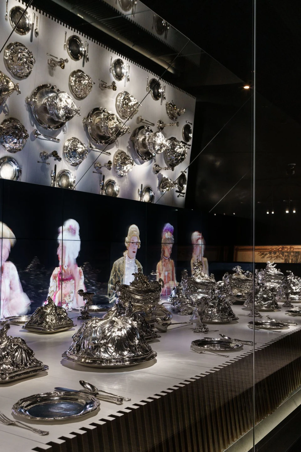 Display case with an assortment of silverware on shelves and walls, and holograms of people in historical attire in the background.