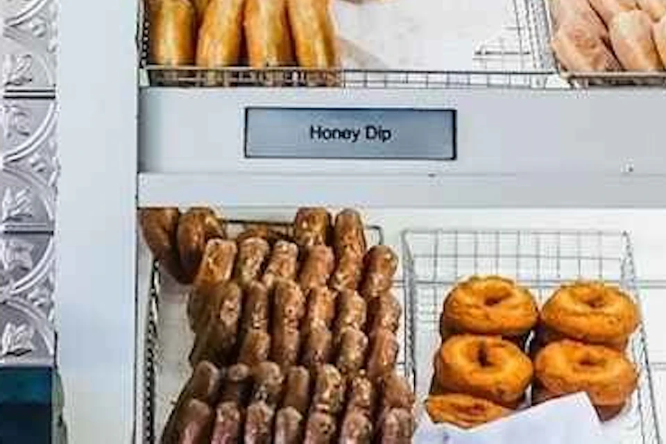 Two employees in a donut shop prepare orders with pastries behind them and filled paper bags on the counter.
