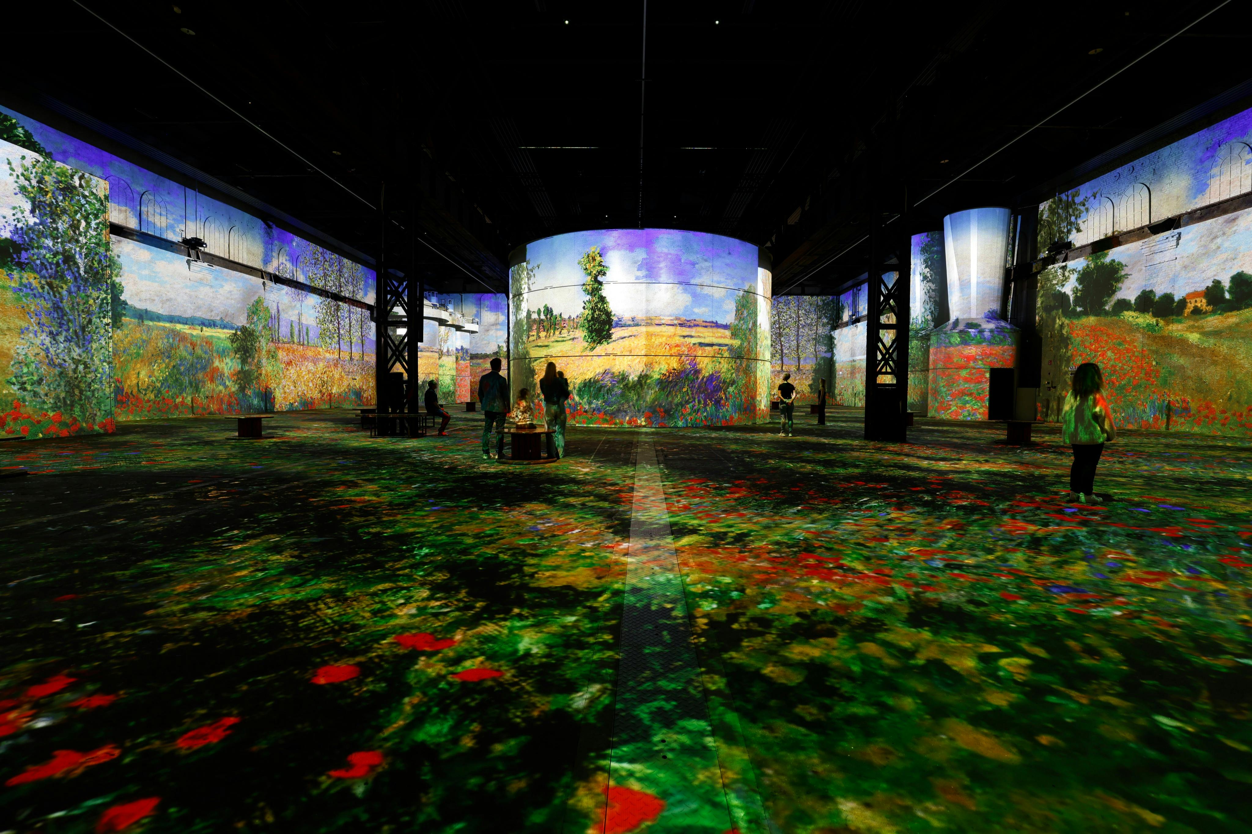 Άνθρωποι περπατούν μέσα από μια καθηλωτική εικαστική εγκατάσταση με προβολές πολύχρωμων, ιμπρεσιονιστικών τοπίων σε τοίχους και δάπεδα.