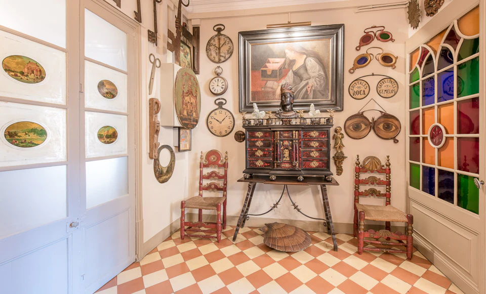 Room with checkered floor, ornate chairs, a decorated cabinet, and walls adorned with antique clocks, paintings, and other vintage items.