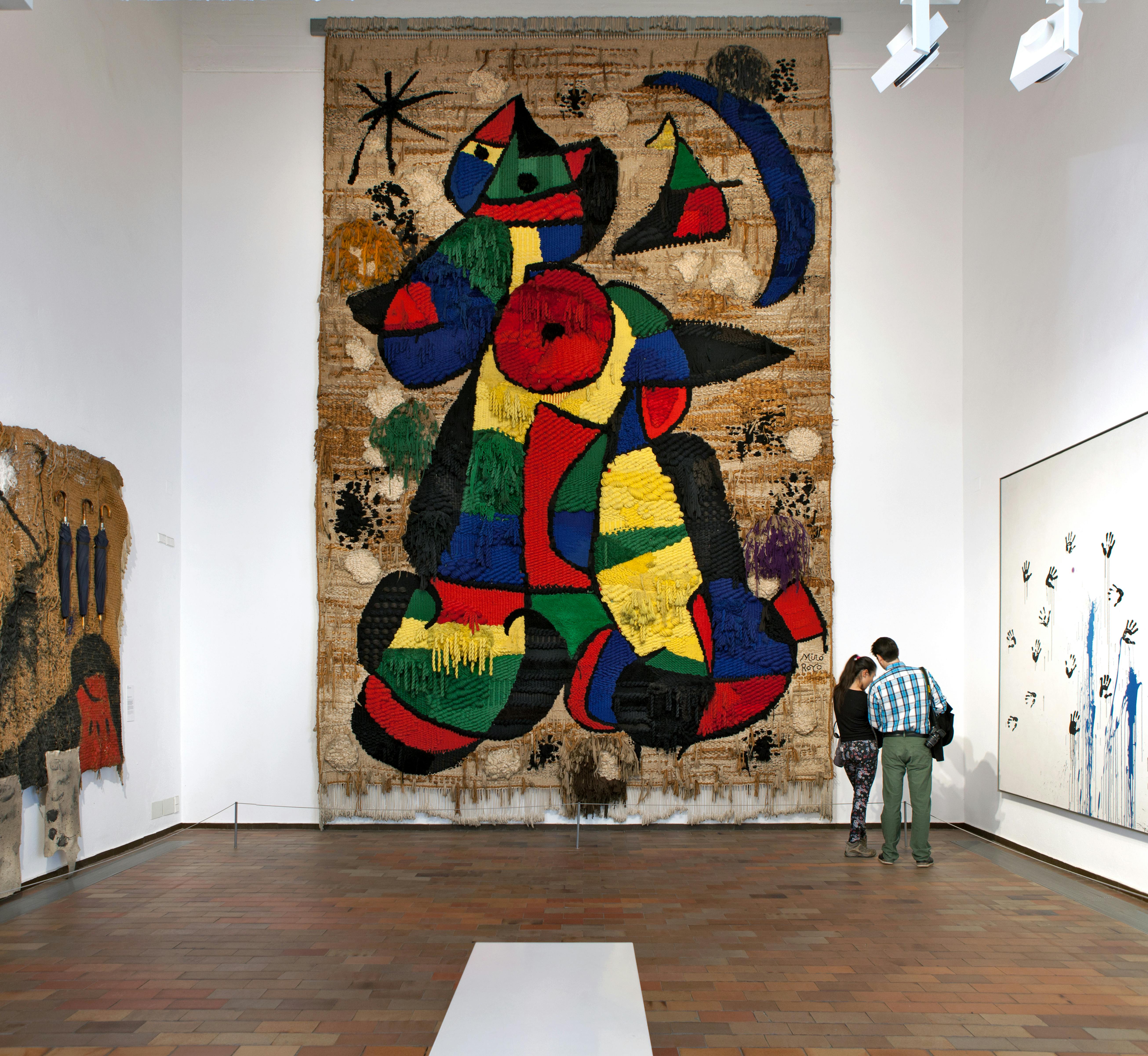 Two people observe a large, colorful abstract tapestry in an art gallery with white walls and wooden floor.