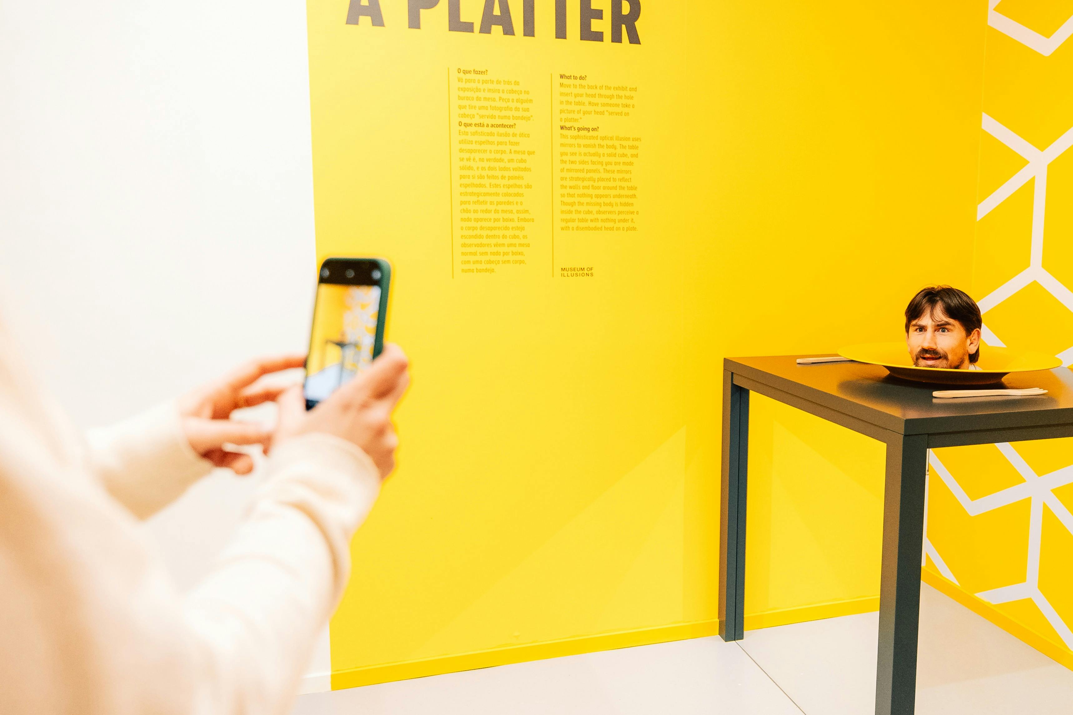 A man's head appears on a plate on a black table in a yellow room. Another person is taking a photo with a phone.