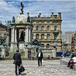 Ett offentligt torg med ett monument, människor som sitter på trappor och två män i kostymer som går. Historiska byggnader i bakgrunden.