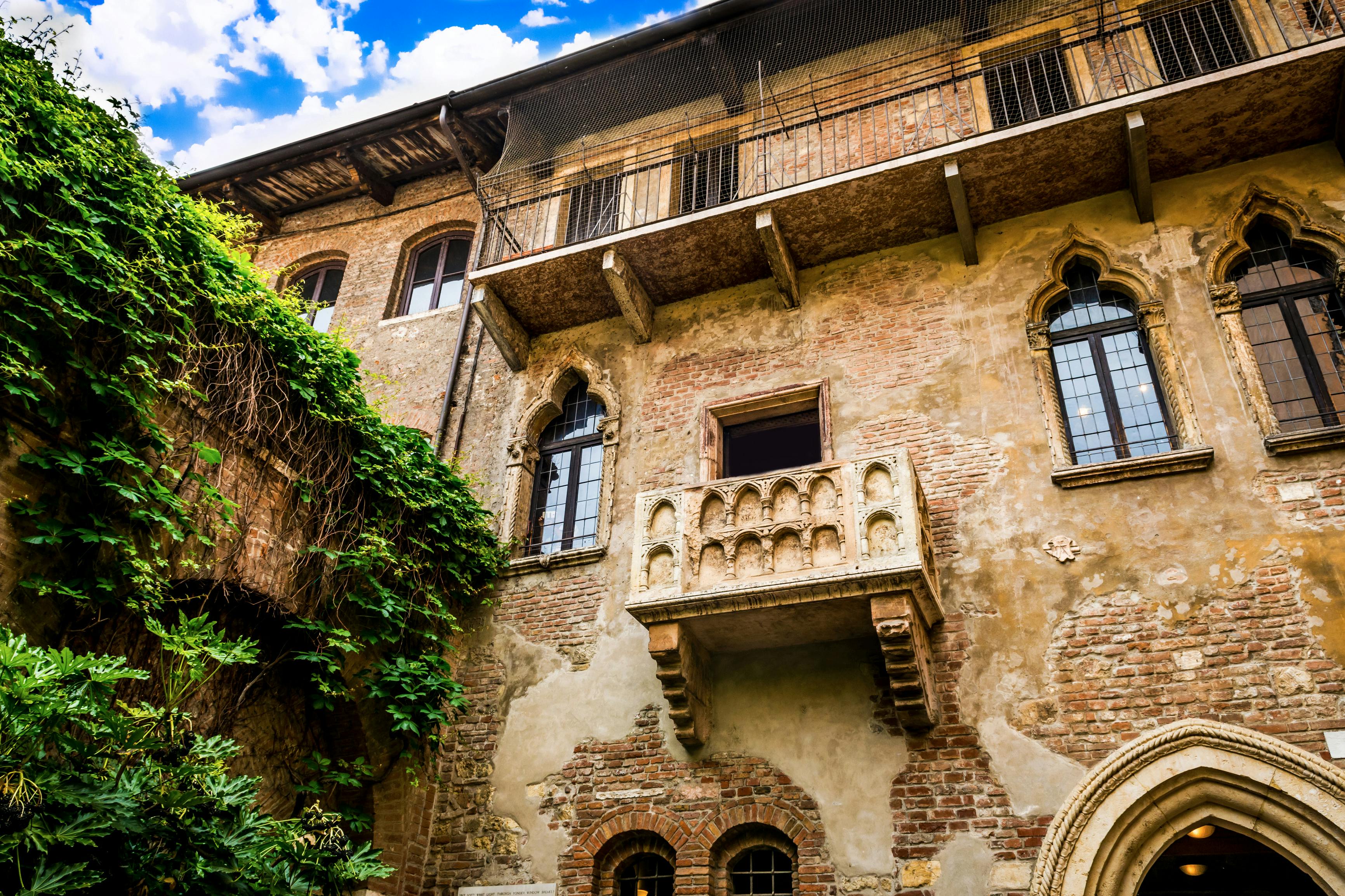 A historic brick building with a stone balcony, arched windows, and ivy-covered walls under a blue sky with clouds.