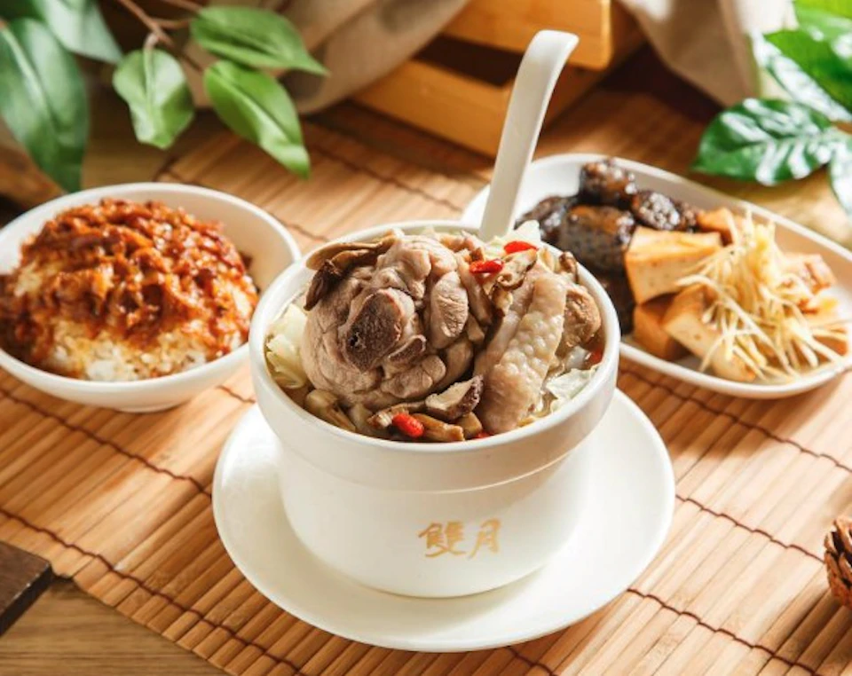 A bowl of soup with meat and vegetables, accompanied by two side dishes, placed on a bamboo mat with a leafy background.