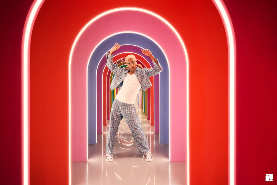 A person posing in a vibrant hallway with illuminated red and pink arches, wearing patterned clothing and sneakers.