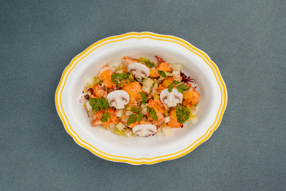 A dish with lobster pieces, sliced mushrooms, diced vegetables, and herbs, served on a white plate with yellow trim.