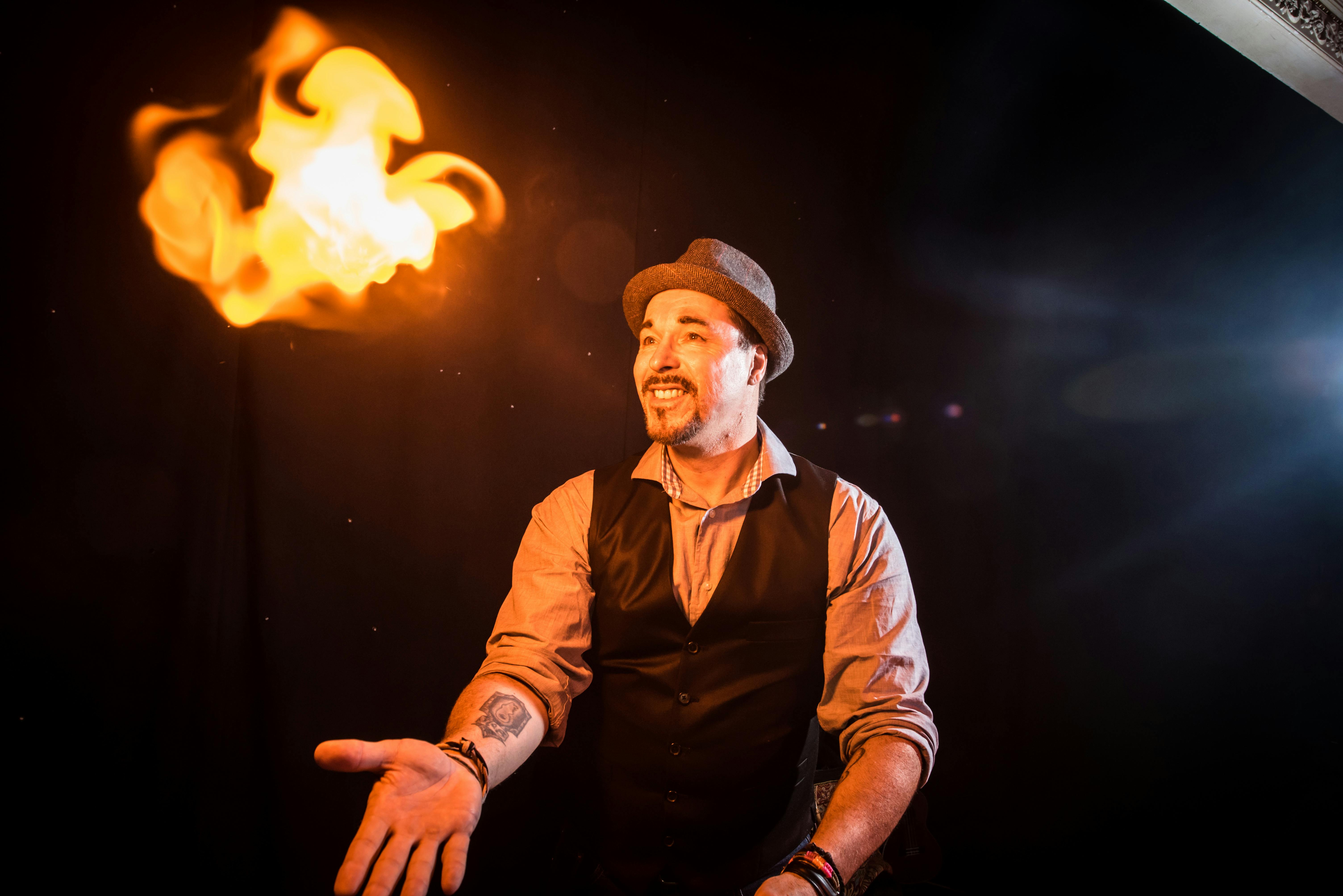 一个戴着帽子和背心的男人站在黑暗的房间里微笑着，伸手拿着一团火球。